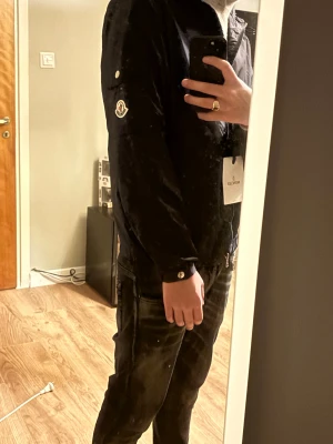 Svart vindjacka från Moncler - Säljer en svart vindjacka från Moncler med huva och snygga detaljer som dragkedja och knappar. Jackan har Moncler-loggan på ärmen och en clean, sportig look. Perfekt för dig som gillar stilrena och funktionella plagg. Passar dig som vill ha en jacka med cool vibe.