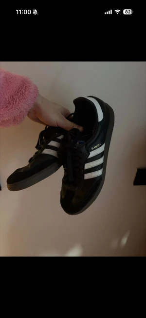 Adidas Samba svarta sneakers - Klassiska Adidas Samba sneakers i svart skinn med vita detaljer och de ikoniska tre ränderna på sidorna. Låg modell med snörning och gummisula. Perfekta för dig som gillar retrostil och streetwear.
