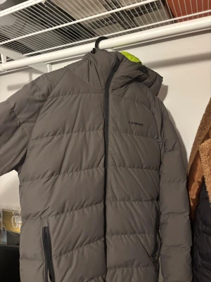 Grå pufferjacka från Everest - Grå pufferjacka från Everest med huva och svarta dragkedjor. Jackan har ett stilrent, quiltat mönster och är perfekt för kalla dagar. Fodret har en touch av limegrönt som syns vid huvan. Snygg och funktionell design för dig som gillar att hålla värmen.