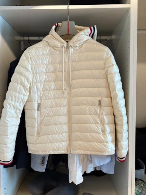 Vit Moncler dunjacka med huva - Säljer en snygg vit dunjacka från Moncler med huva och dragkedja framtill. Jackan har quiltad design, två fickor med dragkedja och coola röd-blå-vita detaljer på huva och ärmslut. Perfekt för dig som vill ha en stilren och sportig look.