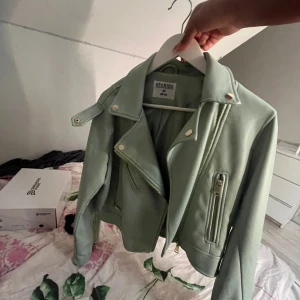 Mintgrön skinnjacka från & Other Stories - Snygg mintgrön skinnjacka från & Other Stories i bikerstil. Jackan har silvriga dragkedjor, klassisk krage med tryckknappar och coola detaljer vid axlarna. Perfekt för dig som vill sticka ut med en fräsch färg och edgy vibe.