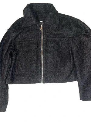 Svart teddyjacka med dragkedja - Svart teddyjacka med croppad passform och två stora fickor framtill. Jackan har en grov dragkedja och mjukt, fluffigt material som ger en cool vibe. Perfekt för dig som gillar att sticka ut med en enkel men trendig look. 