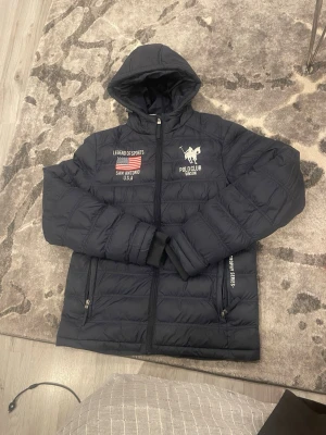 Svart pufferjacka från Vinson Polo Club - Svart pufferjacka med huva från Vinson Polo Club. Jackan har broderade detaljer med amerikansk flagga och text, samt logga på bröstet. Quiltad design, dragkedja framtill och fickor med dragkedja. Perfekt för kalla dagar och har en sportig vibe.