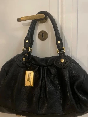 Svart handväska från Marc Jacobs - Säljer en snygg svart handväska från Marc Jacobs med guldfärgade metalldetaljer och en cool metallbricka framtill. Väskan är tillverkad i skinn och har dubbla handtag med dekorativa nitar och ringar. Perfekt för dig som gillar stilrena accessoarer med lyxig känsla.