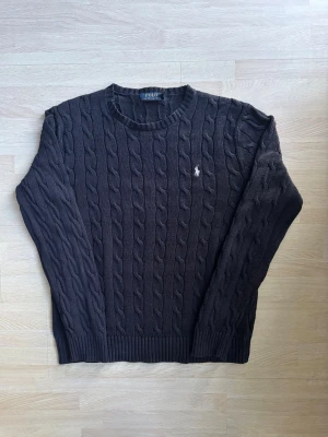 Mörkblå stickad tröja Polo Ralph Lauren - Snygg mörkblå stickad tröja från Polo Ralph Lauren i klassisk kabelstickad design. Tröjan har rund hals, ribbade muddar och den ikoniska broderade loggan på bröstet. Perfekt för dig som gillar stilrena och tidlösa plagg.