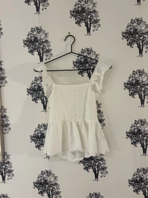 Vit blus med volang från SHEIN - Superfin vit blus från SHEIN i storlek XS. Blusen har breda volangärmar, fyrkantig halsringning och broderade detaljer. Den är croppad med en volangkant nedtill och har snörning i ryggen. Materialet är 95% polyester och 5% elastan.