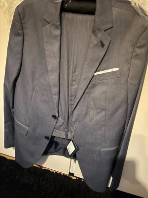 Blå kavaj med klassisk passform - Snygg blå kavaj med två knappar framtill och klassiska slag. Kavajen har en bröstficka med vit näsduk och två fickor med lock. Materialet ser ut att vara slätt och lätt, perfekt för en stilren look.