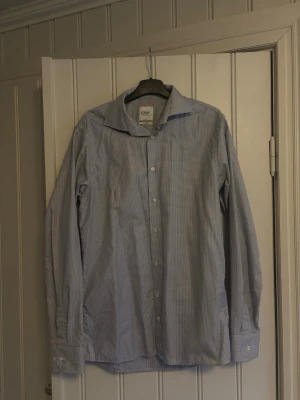 Oscar of Sweden slim fit skjorta - Snygg slim fit skjorta från Oscar of Sweden i vitt och blått småmönstrat tyg. Skjortan har klassisk krage, långa ärmar och knäppning framtill. Perfekt för dig som gillar stilrena detaljer och en smalare passform.