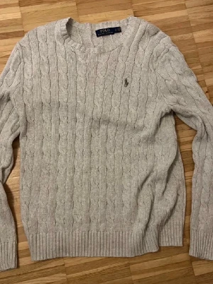 Ljusgrå kabelstickad tröja Polo Ralph Lauren - Säljer en ljusgrå kabelstickad tröja från Polo Ralph Lauren i storlek S. Tröjan har rund hals, lång ärm och klassisk broderad logga på bröstet. Tillverkad i 100% bomull med ribbade muddar vid ärmslut och nederkant. Perfekt för chill höst- och vinterstil.