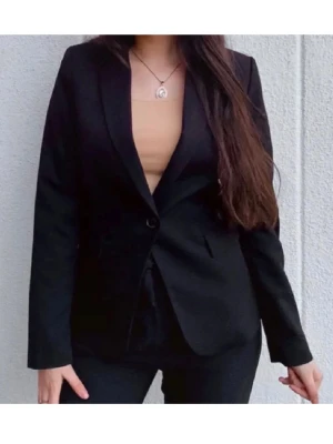 Svart kavaj från Mango - Stilren svart kavaj från Mango med klassisk krage och enkel knappstängning framtill. Kavajen har två fickor med lock och är figursydd för en snygg passform. Perfekt att matcha med kostymbyxor eller jeans för en cool look.