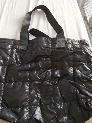 Svart quiltad shoppingväska - Stor och rymlig svart shoppingväska med quiltad design och blanka ytor. Väskan har breda handtag i tyg och stängs med dragkedja. Insidan har ett fack för enkel organisering av dina grejer. Perfekt för att bära med allt du behöver under dagen.