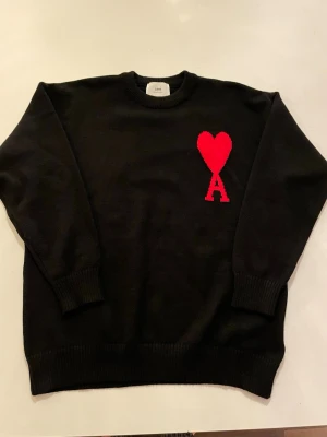 Svart stickad tröja från Ami Paris - Svart stickad tröja från Ami Alexandre Mattiussi med rund hals och ribbade muddar. På bröstet finns ett stort rött hjärta med bokstaven A under, vilket ger en unik och cool detalj. Perfekt för dig som gillar stilrena plagg med en twist. Fick den i julklapp men passade inte riktigt min smak därför säljer jag den!