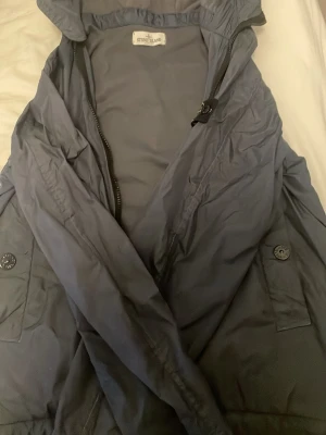 Blå vindjacka från Stone Island - Säljer en blå vindjacka från Stone Island med huva och dragkedja. Jackan har två fickor med knappstängning och en stilren design. Perfekt för dig som vill ha en lätt och snygg jacka till vardags. Passar till många olika outfits.