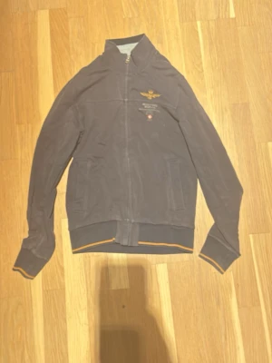 Mörkblå ziptröja Aeronautica Militare - Säljer en mörkblå ziptröja från Aeronautica Militare med broderad logga och text på bröstet. Tröjan har ribbade muddar med orange detalj, hög krage och dragkedja framtill. Perfekt för dig som gillar sportig och stilren look.