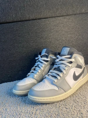Nike Air Jordan 1 Mid grå/vit - Nike Air Jordan 1 Mid sneakers i grått och vitt med klassisk svart swoosh på sidan. Skorna har snörning, rund tå och platt sula. Tillverkade i skinn och syntetmaterial, med ikonisk Air Jordan-logga på sidan och tungan. Perfekt för dig som gillar streetwear och sneakers.