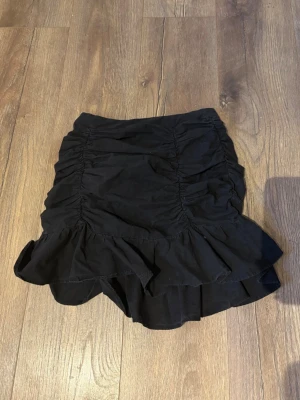 Svart volangkjol från SHEIN - Säljer en svart kortkjol från SHEIN i storlek XS. Kjolen har snygga rynkade detaljer längs sidorna och en bred volangkant nedtill som ger en cool och feminin vibe. Perfekt för dig som gillar trendiga och stilrena plagg. Materialet känns lätt och bekvämt.