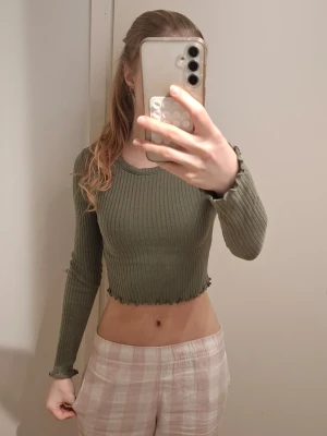 croppad ribbad tröja - Säljer en olivgrön croppad långärmad topp från FB Sister i ribbad bomull. Toppen har vågig kant nertill och vid ärmslut, samt en tight passform som framhäver figuren. Perfekt till jeans eller kjol för en trendig look.