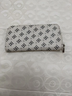 Vit plånbok från Don Donna - Snygg vit plånbok från Don Donna med svart logomönster över hela ytan. Plånboken har dragkedja runtom och flera fack för kort, sedlar och mynt. 