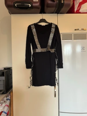 Svart kortklänning Ida Sjöstedt - Svart långärmad klänning från Ida Sjöstedt i storlek S. Klänningen har silvriga detaljer med nätmönster över axlar och midja samt långa band som hänger ner. Materialet är mestadels bomull med lite elastan för stretch. Snygg och unik design med coola kontraster. Den ör i fint skick, men jag tycker att glittret ramlar av lätt. 