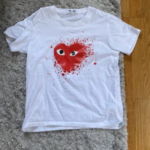Vit t-shirt Comme des Garçons Play S - Säljer en vit t-shirt från Comme des Garçons Play i storlek S. Framsidan har ett stort rött hjärta med ögon och coolt splattertryck. Klassisk rund hals och korta ärmar. Materialet är mjuk bomull som känns skönt mot huden. Små sprickor i trycket som ni ser på bilderna
