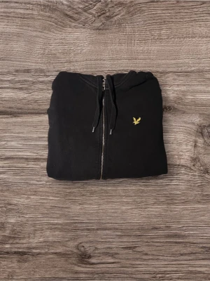 Svart Lyle & Scott zip - Svart Lyle & Scott zip-hoodie i klassisk design. Hel dragkedja, huva med dragsko och diskret broderad örnlogga på bröstet. Skönt och mjukt material med bra passform – perfekt till vardags. I fint skick.