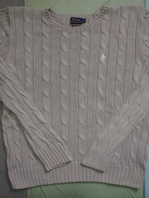 Stickad tröja Polo Ralph Lauren - Säljer en ljusgrå stickad tröja från Polo Ralph Lauren med klassiskt kabelstickat mönster och rund hals. Tröjan har långa ärmar och en broderad vit logga på bröstet. Perfekt för dig som gillar stilrena och tidlösa plagg.