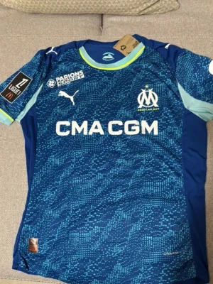 Blå Marseille fotbollströja Puma - Säljer en blå fotbollströja från Puma med Olympique de Marseille-logga på bröstet och digitalt mönster i ljusblått. Tröjan har rund hals med gul kant och flera sponsortryck. Perfekt för dig som älskar fotboll och vill representera ditt lag.