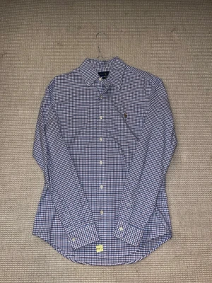 Rutig skjorta från Ralph Lauren - Säljer en slim fit skjorta från Ralph Lauren med blått och vitt rutmönster. Skjortan har klassisk krage, knappar framtill och lång ärm. Materialet är stretchig bomull som är skönt mot huden. Ikonisk broderad logga på bröstet för extra stilpoäng.