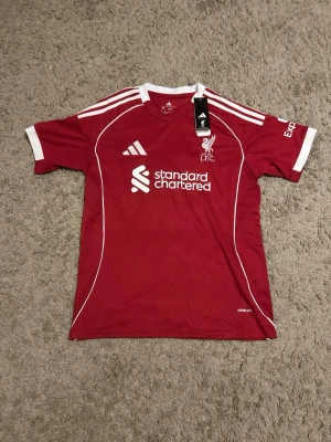 Liverpool FC adidas matchtröja röd - Liverpool FC matchtröja från adidas i röd med vita detaljer och broderat klubbmärke på bröstet. Tröjan har vita ränder på axlarna, vit logga och sponsortryck framtill. Tillverkad i lätt polyester med Aeroready-teknologi för extra komfort.