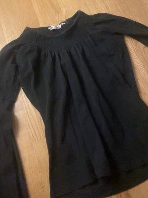 Svart långärmad topp från Instinct - En svart långärmad topp från Instinct i storlek S. Toppen har en rundad halsringning med smockdetaljer upptill som ger en snygg rynkad effekt. Passformen är normal och tyget känns mjukt och följsamt, perfekt till många olika looks.