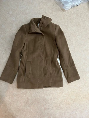 Brun kappa från H&M med hög krage - Snygg brun kappa från H&M i storlek 36 med hög krage och dekorativa knappar. Jackan har två fickor framtill och är tillverkad i ett mjukt ullblandat material. 