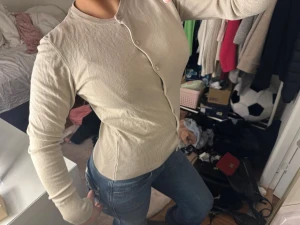 Beige kofta med sned knäppning - Säljer en beige kofta med lång ärm och unik sned knäppning framtill. Tröjan är figurnära med ribbade muddar och nederkant. Perfekt att styla med jeans för en clean look. Obs nopprig