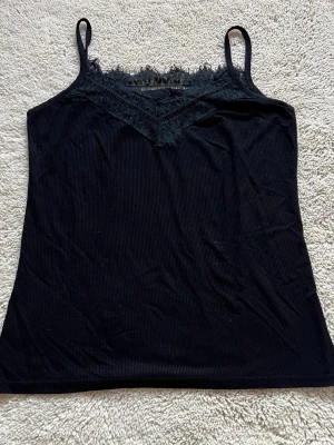 Svart ribbad linnetopp  - Säljer en svart ribbad linnetopp med smala axelband och snygg spetsdetalj vid halsringningen. Toppen har en v-ringad front och ärmlös design. Storleken är både för Xs och S. 