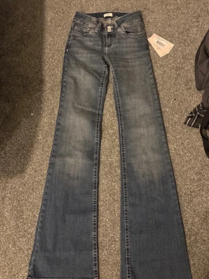 Bootcut jeans från Nelly, mörkblå - Bootcut jeans från Nelly i mörkblå tvätt med låg midja. Snygga detaljer med vit zigzag-söm på bakfickorna och klassisk femficksdesign. Jeansen har breda benslut och är gjorda i jeansmaterial med en cool faded effekt framtill.