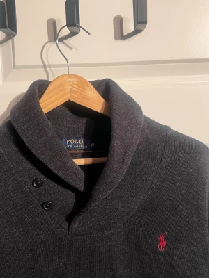 Mörkgrå kofta från Polo Ralph Lauren - Säljer en mörkgrå kofta från Polo Ralph Lauren med snygg krage och två knappar vid halsen. Tröjan har bruna mockadetaljer på armbågarna och den klassiska röda loggan broderad på bröstet. Perfekt för dig som gillar stilrena och tidlösa plagg.