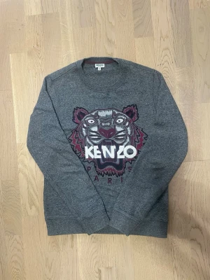 Grå Kenzo sweatshirt med tiger - Säljer en grå sweatshirt från Kenzo i storlek XS med det ikoniska broderade tigermotivet och texten 'KENZO PARIS' framtill. Tröjan har rund halsringning, långa ärmar och är gjord i mjuk bomullsmix. Hör av dig vid intresse, pris kan diskuteras!