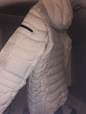 Vit Peak Performance pufferjacka - Säljer en vit pufferjacka från Peak Performance med huva och svarta detaljer. Jackan har dragkedja framtill, ficka med dragkedja på ärmen och justerbar huva. Perfekt för kalla dagar och har en snygg, quiltad look. Nypris 3700kr