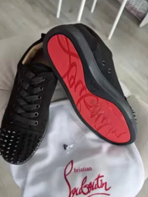Svarta sneakers från Christian Louboutin - Säljer ett par svarta sneakers från Christian Louboutin med ikonisk röd sula och svarta nitar på tån. Skorna har snörning och är tillverkade i mocka med detaljer i skinn. Perfekta för dig som vill sticka ut med en edgy och lyxig stil.