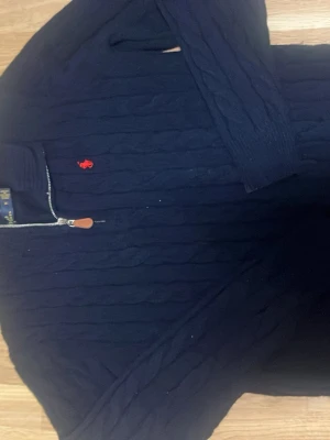 Marinblå stickad tröja Polo Ralph Lauren - Säljer en marinblå stickad tröja från Polo Ralph Lauren med halv dragkedja och klassisk röd broderad logga på bröstet. Tröjan har lång ärm, ribbade muddar och är perfekt för kyligare dagar.