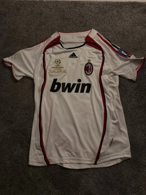 AC Milan Champions League-tröja - AC Milan Champions League-tröja från finalen 2007 med Maldini och nummer 3 på ryggen, tillverkad av Adidas. Tröjan är vit med röda och svarta detaljer, broderade märken och tryck. 