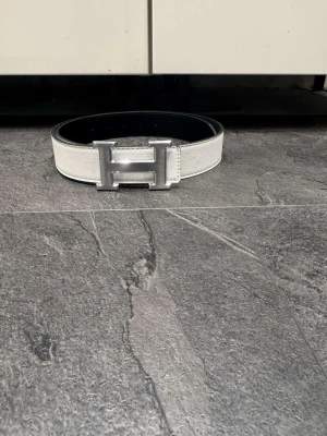 Vitt hermees bälte med silver logga - Snyggt skärp i vitt och svart med ett stort metalliskt H-spänne framtill. Skärpet är tillverkat i skinn och har en stilren, klassisk design som sticker ut. Perfekt för att ge outfiten en extra touch. Passar både jeans och kostymbyxor.