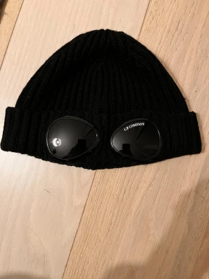 Svart C.P. Company mössa med goggles - Säljer en svart ribbstickad mössa från C.P. Company med ikoniska goggles på uppviket. Perfekt för det kalla vädret. OBS, reser iväg den 26:e December och kommer tillbaka den 7:e Januari. Skriv gärna privat vid fler frågor. 100% äkta