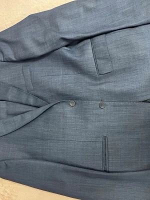 Grå kavaj från Satisini inklusive byxor - Snygg grå kavaj från Satisini med klassisk passform och tre knappar framtill. Kavajen har smala slag, raka fickor och ett diskret mönster i tyget. Perfekt för dig som gillar stilrena och tidlösa plagg.