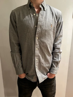Grå skjorta från Ralph Lauren - Snygg grå skjorta från Ralph Lauren med klassisk button-down krage och broderad logga på bröstet. Skjortan har lång ärm, vita knappar och är gjord i mjuk bomull. Perfekt till jeans och sneakers för en chill look.