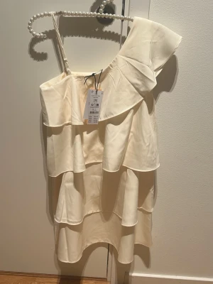 Champagnefärgad omlottklänning  - Säljer en champagnefärgad kortklänning med volanger och omlottdetalj. Klänningen har oneshoulder-design med ett brett axelparti och är tillverkad i ett mjukt, glansigt material som ger en lyxig känsla. 