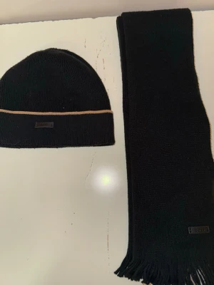 Svart stickad mössa och halsduk från BOSS - Säljer ett set med svart stickad mössa och halsduk från BOSS. Mössan har en beige rand och båda delarna har en snygg BOSS-logga i svart läder. Halsduken har fransar i ändarna och är enkel i designen. Perfekt för kalla dagar.