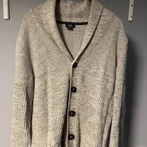 Grå kofta från F&F med knappar - Säljer en grå/ljus beige stickad kofta från F&F i storlek M. Koftan har bred krage, långa ärmar och stängs med fyra svarta knappar framtill. Den är ribbstickad på ärmar och nedre delen, vilket ger en snygg struktur. Perfekt för lager-på-lager under kyliga dagar.