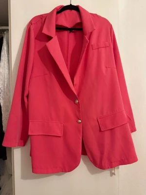 Rosa kavaj med guldknappar - Snygg rosa kavaj med klassisk krage och två guldknappar framtill. Kavajen har två stora fickor med lock och en bröstficka. Passar perfekt för dig som vill sticka ut med färg. Långärmad och med en loose fit.