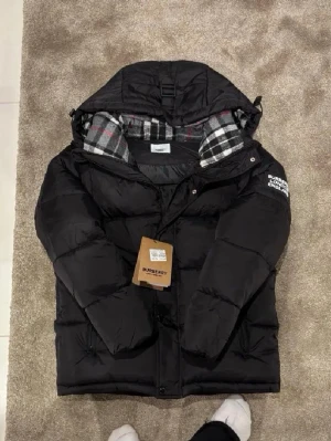 Svart pufferjacka från Burberry - Svart pufferjacka från Burberry med huva och rutig innerfoder i svart, vitt och rött. Jackan har en stor Burberry-patch på ärmen, dragkedja framtill och quiltad design. Perfekt för kalla dagar och har en riktigt schysst streetstil.