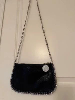 Svart axelväska med kedja - En svart axelväska med glansig finish och silverfärgad kedja som axelrem. Väskan har en rund metallbricka vid dragkedjan och detaljer i metall runt kanterna. Perfekt för dig som gillar en edgy och stilren look.